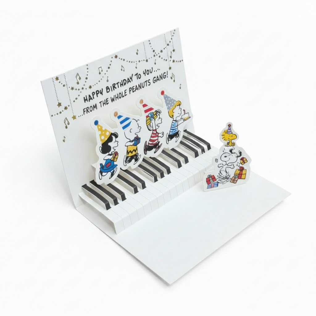A 3D pop-up greeting card featuring cartoon characters playing a piano, displayed open on a wooden surface with a yellow envelope and a cardboard box in the background.