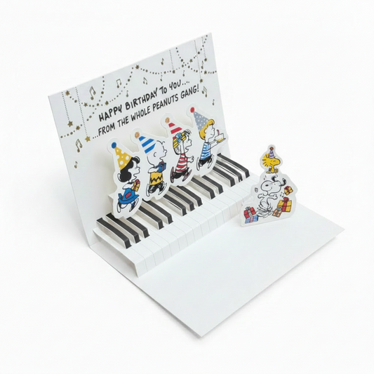 A 3D pop-up greeting card featuring cartoon characters playing a piano, displayed open on a wooden surface with a yellow envelope and a cardboard box in the background.